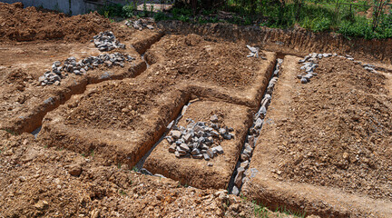An outdoor construction site shows freshly dug trenches filled with rocks and gravel, marking the...