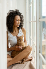 Young woman holding ginger cat and looking out window