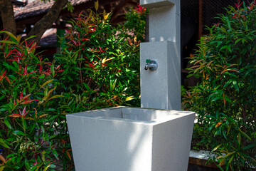 An outdoor sink with a metal faucet surrounded by green and red ornamental plants. The modern stone structure is situated in a shaded garden area, evoking cleanliness and nature.