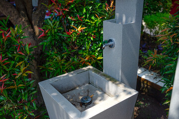An outdoor sink with a metal faucet surrounded by green and red ornamental plants. The modern stone structure is situated in a shaded garden area, evoking cleanliness and nature.