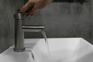 Opening a brushed metal faucet above a pristine white sink, a hand directs a stream of fresh water flowing into the basin, contrasting beautifully with the dark gray tiled wall