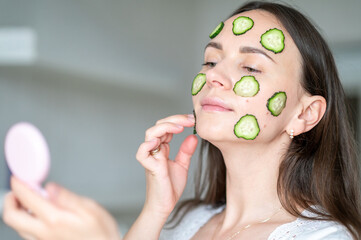 Woman with cucumber slices on her face looks into hand mirror enjoying healthy skin care