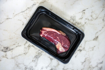 Piece of meat is sitting on a marble countertop. The meat is in a plastic container and he is raw