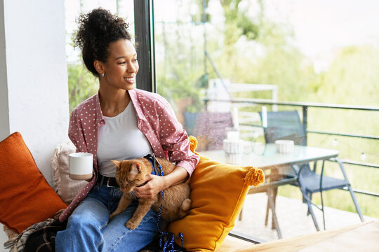 Young woman relaxing at home, holding a ginger cat and drinking coffee - Powered by Adobe
