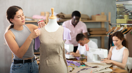 Interested young woman adjusting and pinning fabric on mannequin during hands-on dressmaking class, while classmates sewing and preparing materials at table in background..