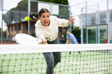 Sporty young girl playing padel on open court on summer day, ready to hit ball with forehand. Sport...