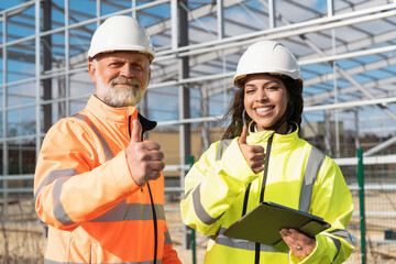 Man and woman construction workers giving thumbs-up as they discuss plans on a site while wearing safety gear and reviewing documents