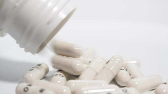 White capsules spill from a plastic pill bottle in slow motion onto a clean white surface. The close-up shot emphasizes the clarity and texture of the capsules.