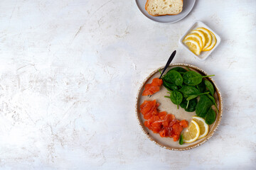 Salted trout fillet thinly sliced ​​on a ceramic plate, served with spinach, lemon and wheat bread. Ingredients for making a sandwich. Healthy eating. Copy space
