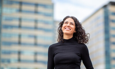 confident woman with curly hair stands outdoors in the city smiling as she looks up