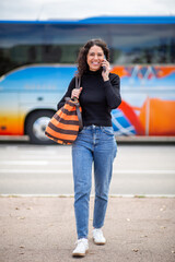 Full body smiling woman talking on phone while walking, with bus in the background