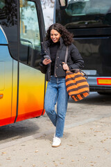 Full body smiling travel woman walking near colorful bus with phone in hand
