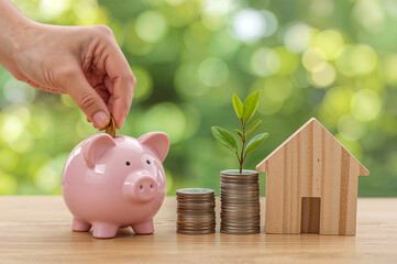 Hand Inserting Coin into Piggy Bank with Wooden House and Coin Plant for Financial Growth Concept