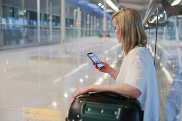 A young woman sits in an airport terminal, checking her phone while resting her arm on a suitcase....