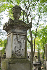 Old stone grave monument with a relief of an angel comforting a sorrowful figure, topped with an urn, in a historic cemetery surrounded by green trees.