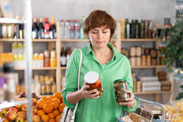 Woman looking at canned vegetables in store. Market visitor choose consider jar with canned tomato...