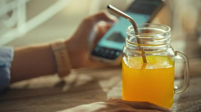A person enjoys a vibrant orange drink from a mason jar while scrolling on a smartphone at a relaxed cafe setting. The warm sunlight creates a cheerful atmosphere perfect for leisurely moments