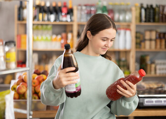 Teen girl hold packages bottles and choose consider juice in store. Market visitor choose consider...