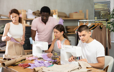 Skillful middle-aged trainer making instructions to trainees, other student making dress detail during sewing lesson
