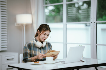 A young woman wearing headphones reads a book peacefully at home, enjoying a quiet study moment in...