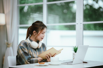 A young woman wearing headphones reads a book peacefully at home, enjoying a quiet study moment in...