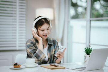A young woman wearing headphones smiles while using her smartphone at home. Surrounded by cozy...