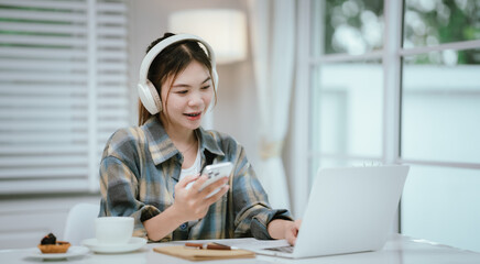 A young woman wearing headphones smiles while using her smartphone at home. Surrounded by cozy...