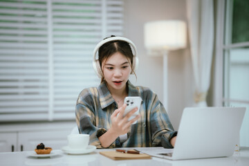 A young woman wearing headphones smiles while using her smartphone at home. Surrounded by cozy...
