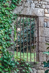 Ancient stone wall with a barred window and ivy in Girona, Spain. Rustic masonry, metal bars, and lush greenery create a charming medieval scene with a garden glimpse beyon