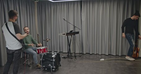 A music band practices in their studio before a concert. The drummer plays the drums while the keyboardist adjusts the keyboard and the guitarist looks on. - Powered by Adobe