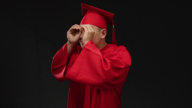 Young chinese man in red graduation cap and gown makes binocular gesture isolated on a black background, symbolizing curiosity and future vision.