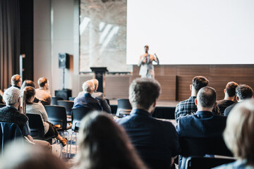 Naklejka na ściany i meble Speaker giving a talk in conference hall at business event. Rear view of unrecognizable people in audience at the conference hall. Business and entrepreneurship concept