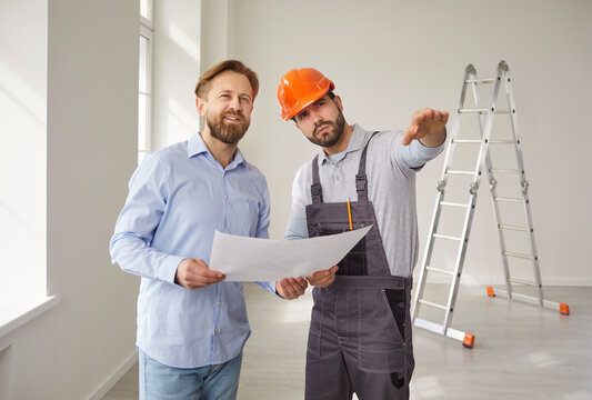 Construction worker in helmet discussing renovation plan with male client at construction site. Professional builder with blueprint talking to architect or engineer, explaining process of changes.