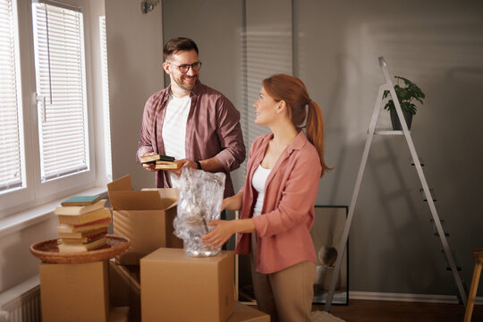 A couple is busy moving into their new home, surrounded by cardboard boxes filled with books and household items, engaging in a joyful and collaborative unpacking experience.