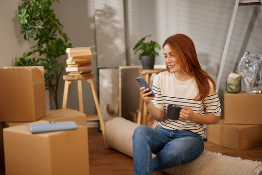 A woman sits on a carpet surrounded by unpacked boxes, sipping coffee from a mug. She smiles while looking at her smartphone, reflecting a relaxed moment during her move. - Powered by Adobe