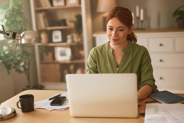 A woman sits at a wooden desk working on her laptop in a bright and inviting home office. Papers are scattered around her while she enjoys coffee, focused on her tasks.
