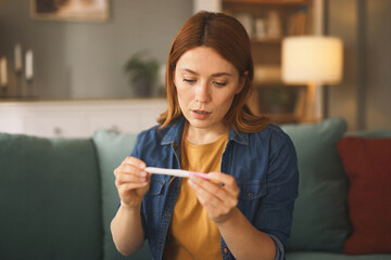 A woman sits on a couch in a comfortable living room, focusing intently on the pregnancy test in...