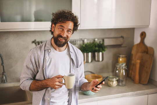 A man with curly hair and a beard holds a coffee mug in one hand while using his phone in a bright and stylish kitchen, creating a cozy morning atmosphere. - Powered by Adobe