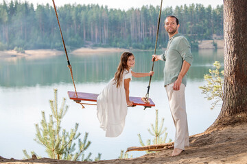 A father swings his daughter on a swing on the shore of a forest lake.