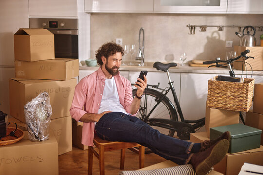 A man sits casually on a chair, using his smartphone while surrounded by unpacked boxes in his new kitchen, enjoying a moment of relaxation on moving day.