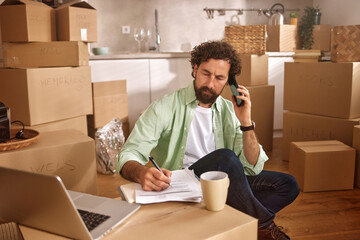 A man sits on the floor of his new home, taking a phone call while writing notes. He is surrounded...