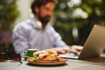 A business professional focuses on tasks on a laptop at an outdoor cafe, enjoying a croissant with ham and vegetables, immersed in a productive morning atmosphere.