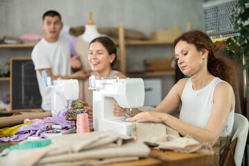 Enthusiastic woman stitching at machine in tailoring studio, enjoying hands-on learning during group sewing class for adults, while fellow learners sharing creative work and friendly conversation