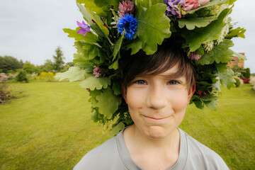 Smiling boy wearing a large floral crown made of oak leaves and wildflowers, standing outdoors on a...