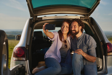 A couple sits in the back of their vehicle, smiling and taking a selfie together. They are surrounded by nature, enjoying a relaxed picnic experience on a beautiful day.