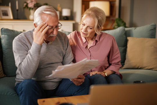 An older Caucasian couple sits on their blue sofa while reviewing financial paperwork and insurance information. They appear concerned while discussing details of the documents in their home.