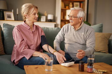 A senior couple is sitting on a couch at home, the woman is checking her blood pressure with a digital monitor, and the man is holding her hand while they look at each other and smile.