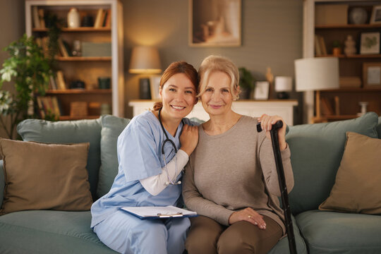 A young, smiling nurse wearing scrubs sits with her arm around a smiling senior woman who is holding a walking stick. Both sit on a teal couch in a living room with bookshelves.
