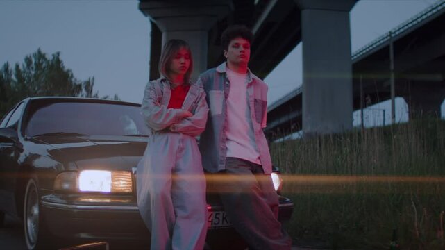 Cool woman and man lean on dark car on roadside near overpass
