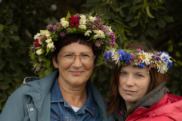 Obraz premium Two women stand close together wearing colorful floral wreaths made of wildflowers and greenery, smiling gently against a leafy natural backdrop.